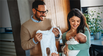 Two people indoors: one holding a white baby onesie, the other cradling an infant wrapped in a white blanket. The setting includ