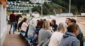 Photo of back's of people eating outdoors sitting at blue benches. Bunting above the seating and beach in the background