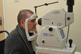 A man sitting on a chair. His chin is resting on a frame, and he is looking forward into a machine called a retinal screening device that looks like a large camera